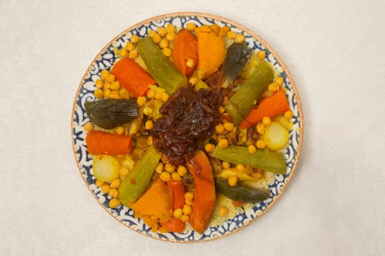 طبق كسكسي مغربي أصيل مقدم في طبق خزفي كبير، مع كومة من الكسكس الأصفر الهش، ولحم ضأن طري محاط بالجزر، البطاطس، الكوسا، والقرنبيط، مع لمسة من اللترنج على الجانب – وجبة تقليدية شهية وغنية بالنكهات.